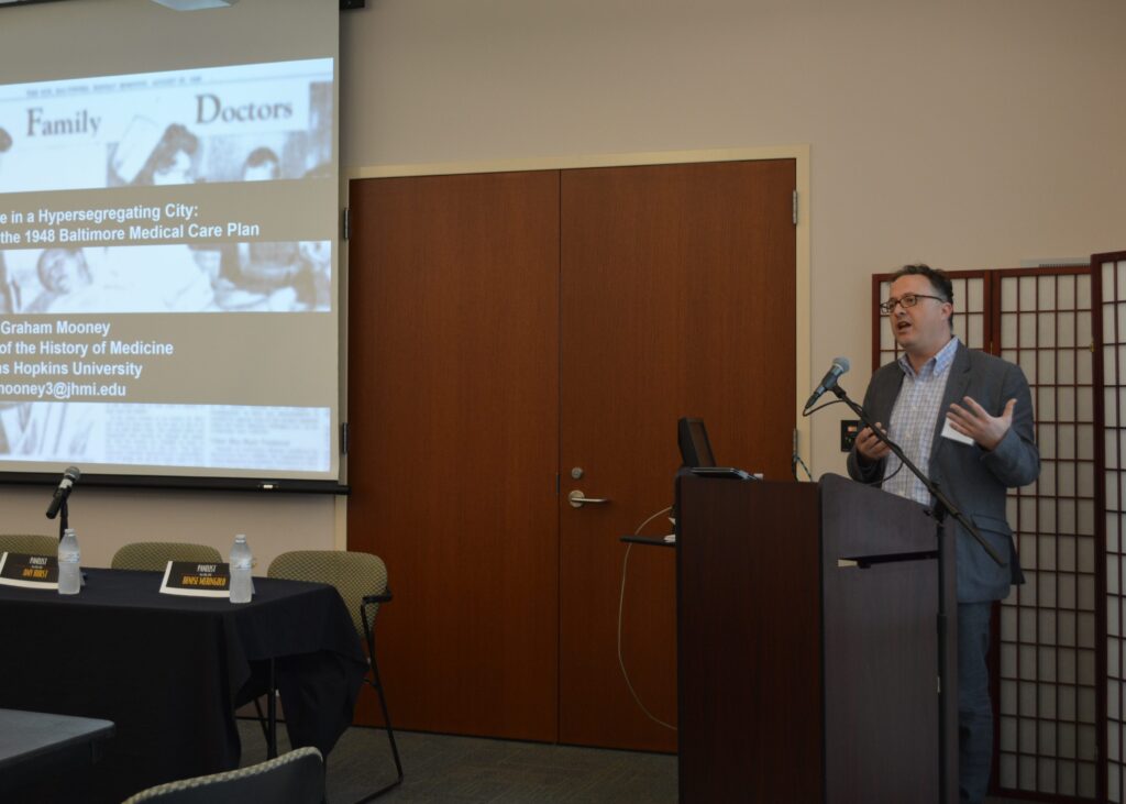 Presenter Graham Mooney stands behind a podium. Presentation slide visible on left side of image.