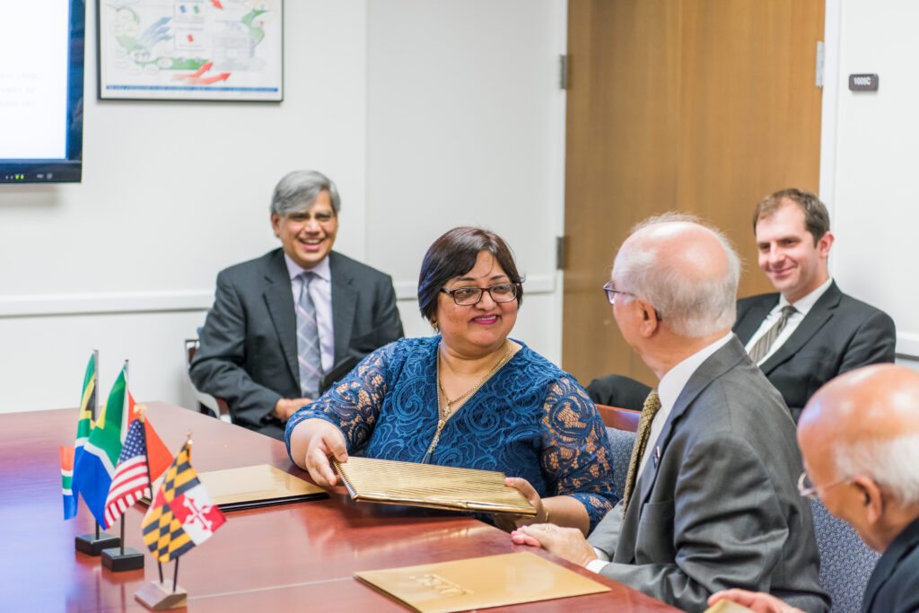 A man and woman in business attire, sitting at a table and smiling, exchange gold folders.