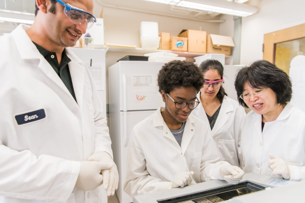 Four scientific researchers gather around a piece of lab equipment, all wearing lab coats and goggles.