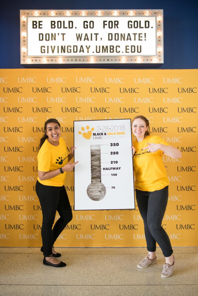 Two young women point to a poster they are holding showing a picture of a thermometer to indicate fundraising level. Above them, a sign reads, "Be bold. Go for gold. Don't wait, donate! givingday.umbc.edu."