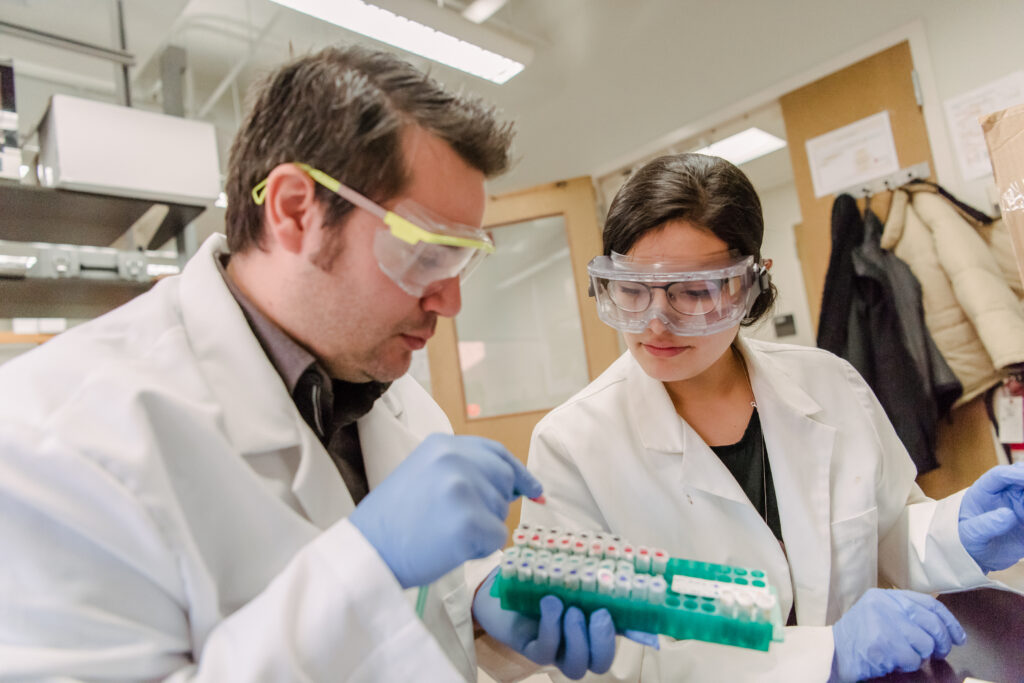 A man and woman wearing lab coats and goggles work in a lab, inspecting samples.