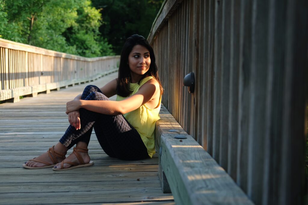 A woman in a yellow shirt, jeans, and sandals sits on a wooden bridge, with trees in the background.