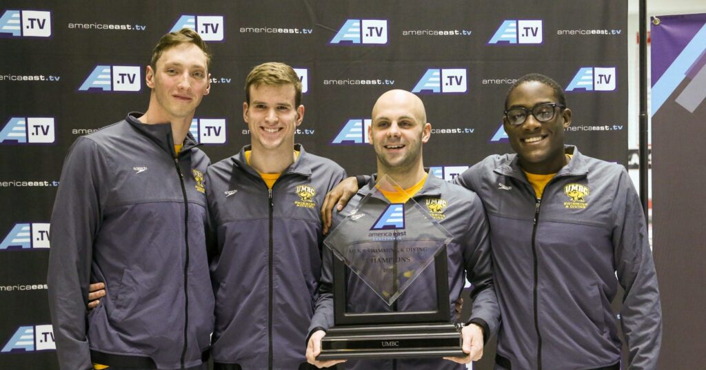 Four male swimmers wearing matching warm-up gear stand in front of an America East sign holding an America East trophy.