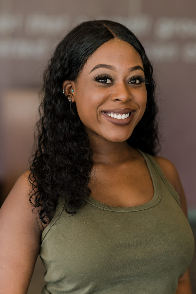 Young woman with long black curly hair, wearing an olive green tank top, smiles at the camera.