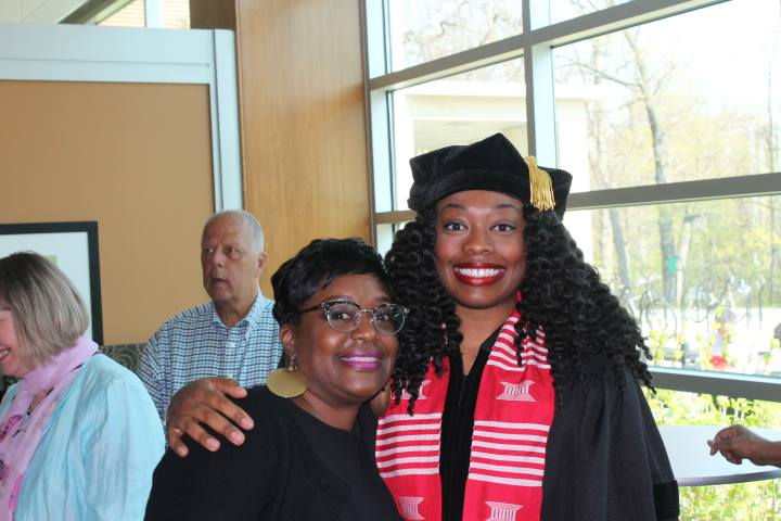A Black woman wearing black cap and gown and red stole with white stripes with her arm around another black woman. Both smile at the camera.