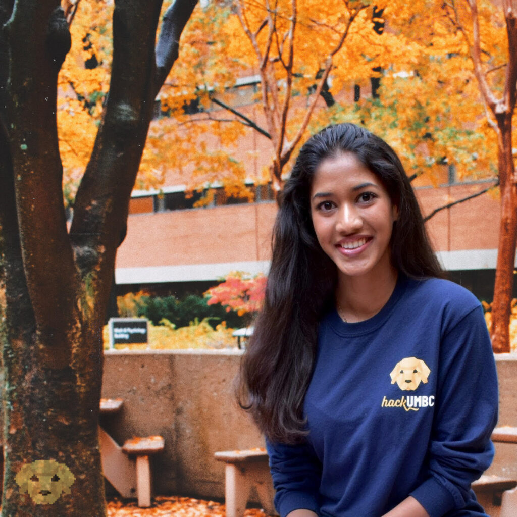 Young woman sits in front of a brick building and tree in the autumn. She wears a long-sleeved t-shirt with a logo for hackUMBC.