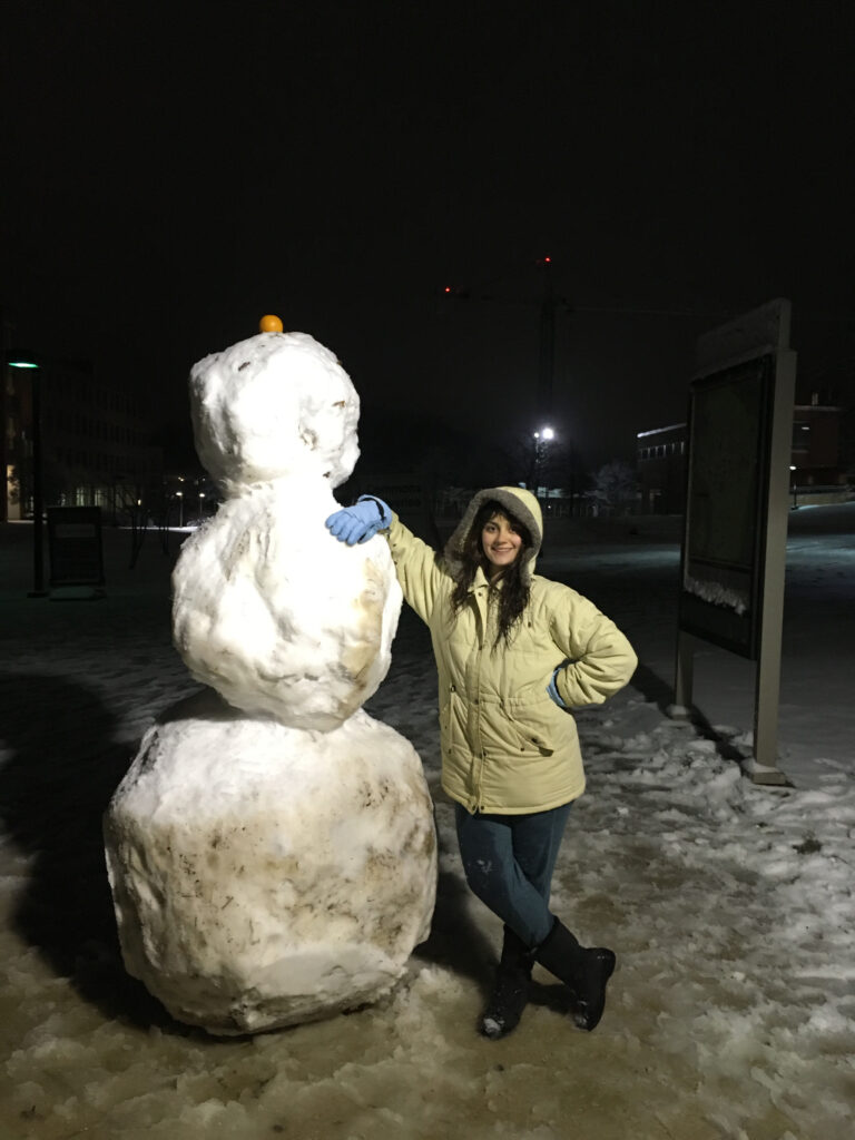 Woman in light green coat and jeans poses next to a very large snowman at night.