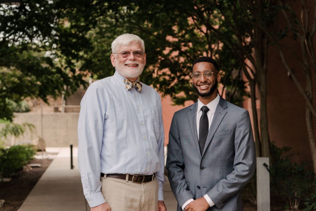 Two men standing on a sidewalk lined by trees. Both men are smiling and wearing glasses. The man on the right is wearing a grey suit, white shirt, and black tie. The man on the left is wearing a light blue shirt, and a bow tie.