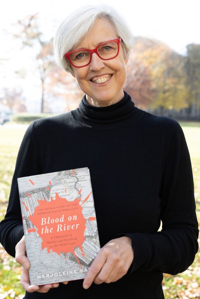 A woman with gray hair wearing a black and red rimmed glasses holds a book with a gray and red cover.