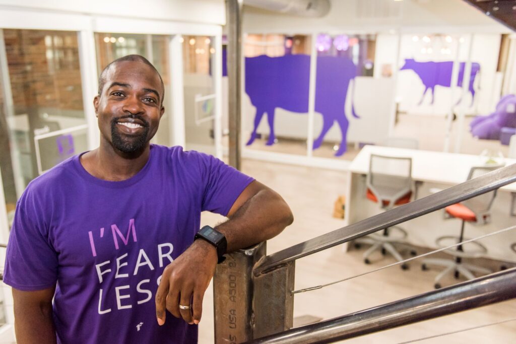 A man wearing a purple t-shirt stands with his arm resting on a bannister. Behind him is a